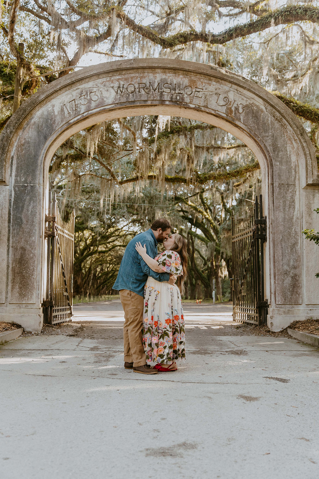 A couple dances on a tree-lined dirt path, with the woman wearing a flowing floral dress and the man in casual attire, surrounded by rustic wooden fences 