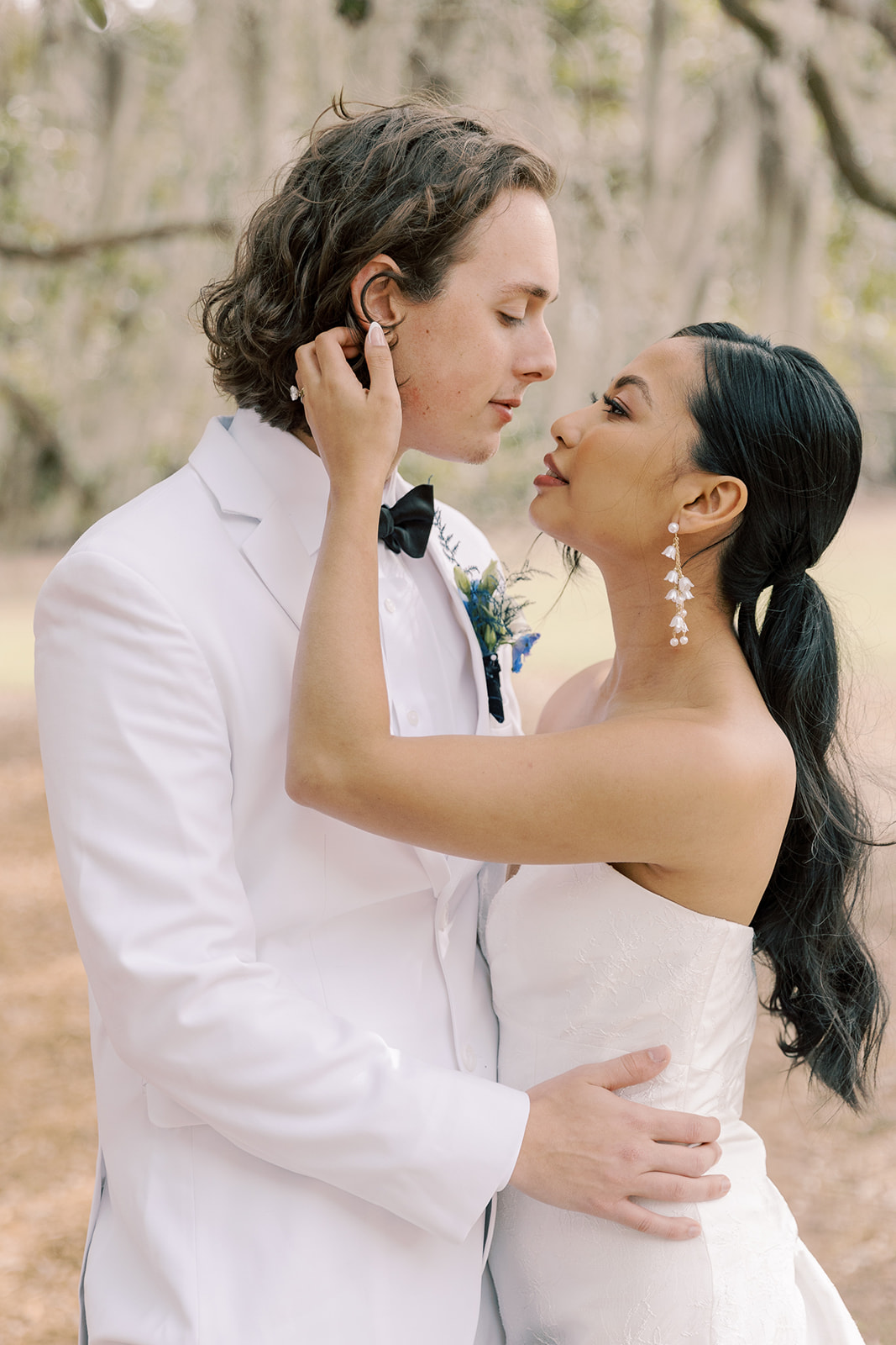 A bride and groom stand close in an outdoor setting. The groom wears a white suit and black bow tie, and the bride is in a strapless white dress, touching his ear. Both have dark hair, and they appear to be about to kiss