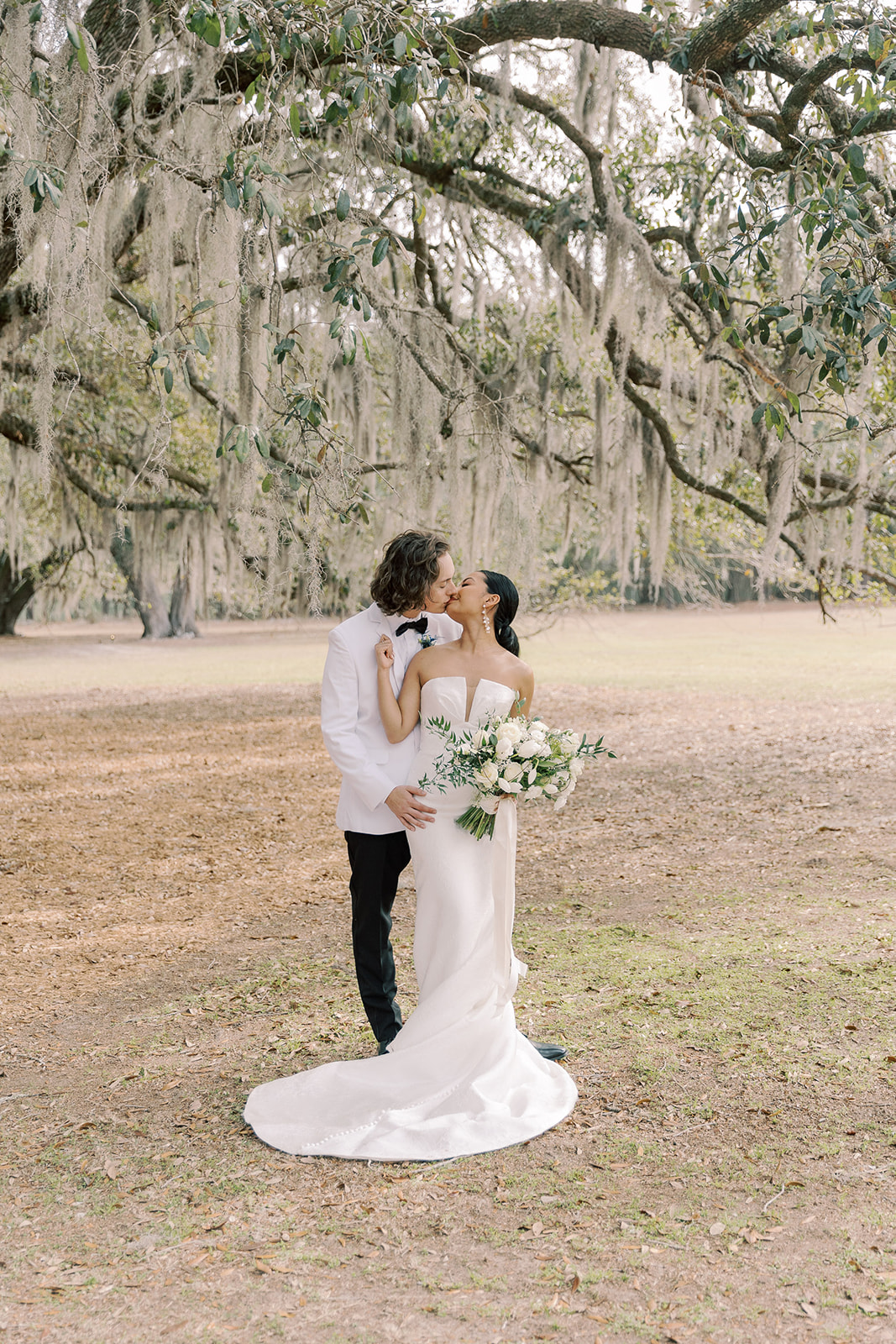 A bride and groom stand close in an outdoor setting. The groom wears a white suit and black bow tie, and the bride is in a strapless white dress, touching his ear. Both have dark hair, and they appear to be about to kiss.