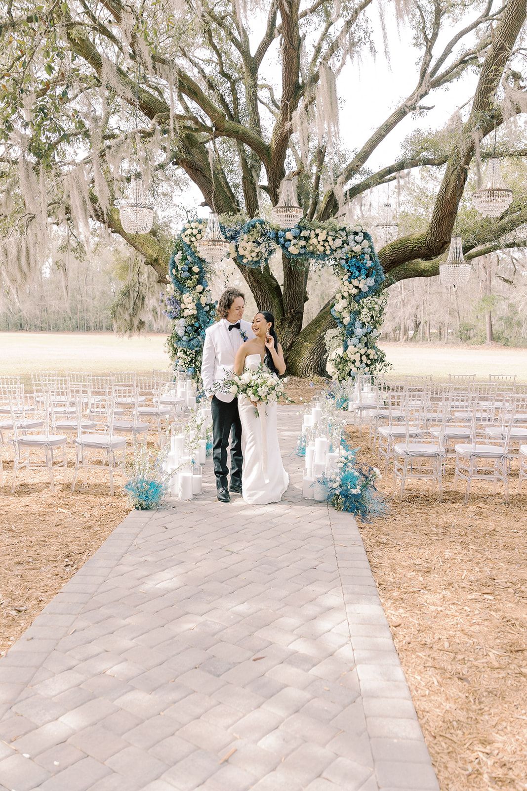 Outdoor wedding setup under a large tree with blue and white floral decorations. Rows of transparent chairs line the aisle, which is adorned with candles and flower arrangements at Hewitt Oaks venue 