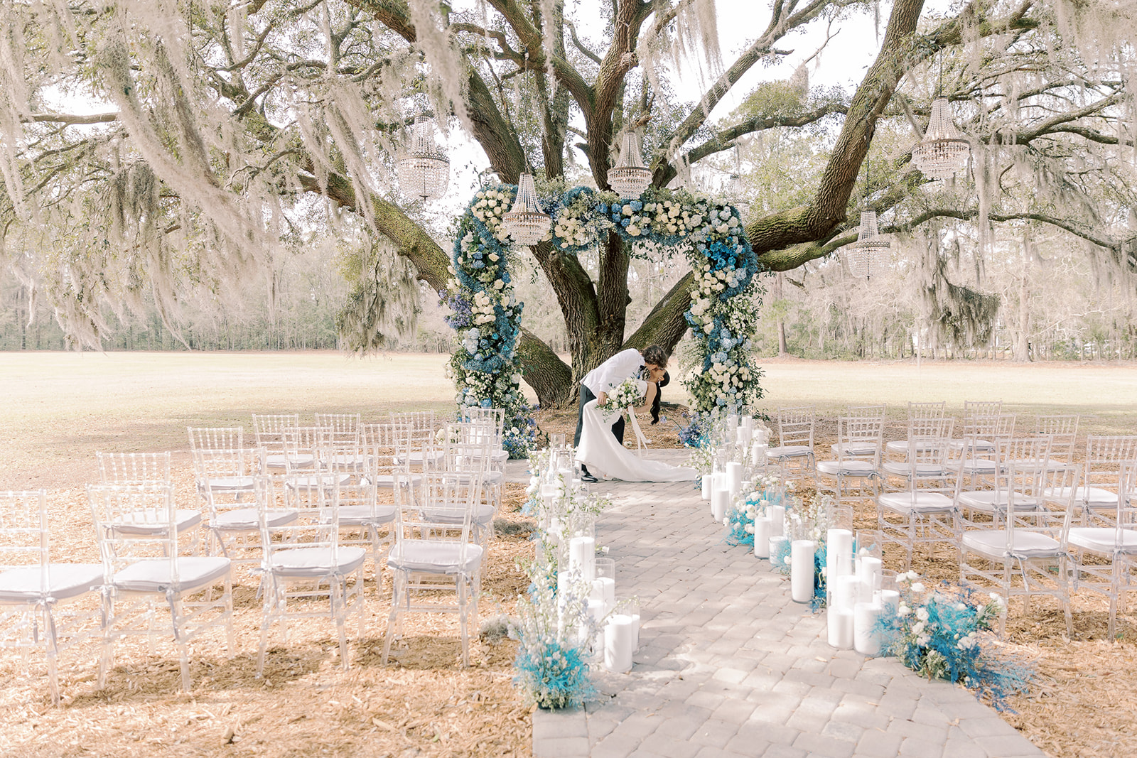 Outdoor wedding setup under a large tree with blue and white floral decorations. Rows of transparent chairs line the aisle, which is adorned with candles and flower arrangements at Hewitt Oaks venue 