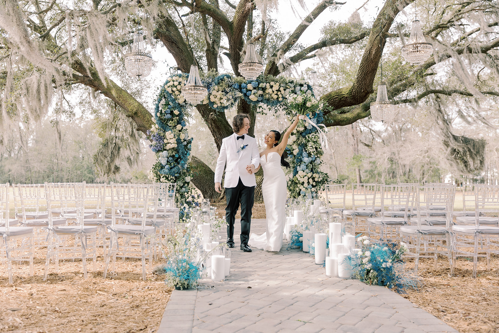 Outdoor wedding setup under a large tree with blue and white floral decorations. Rows of transparent chairs line the aisle, which is adorned with candles and flower arrangements at Hewitt Oaks venue 