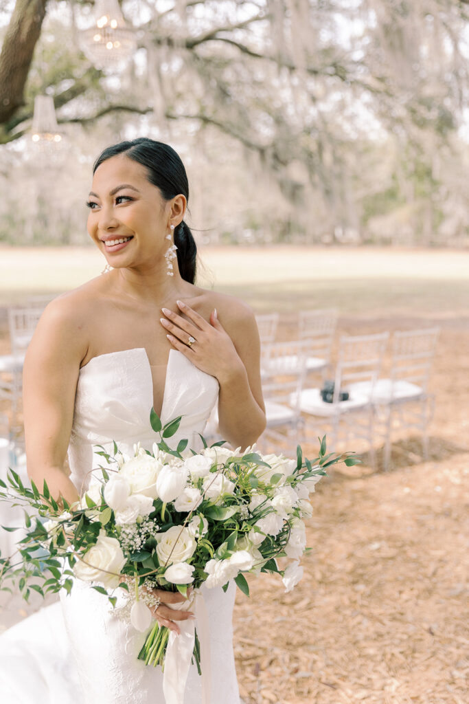 A woman in a white strapless wedding dress holds a bouquet of white flowers outdoors, smiling and looking to the side.