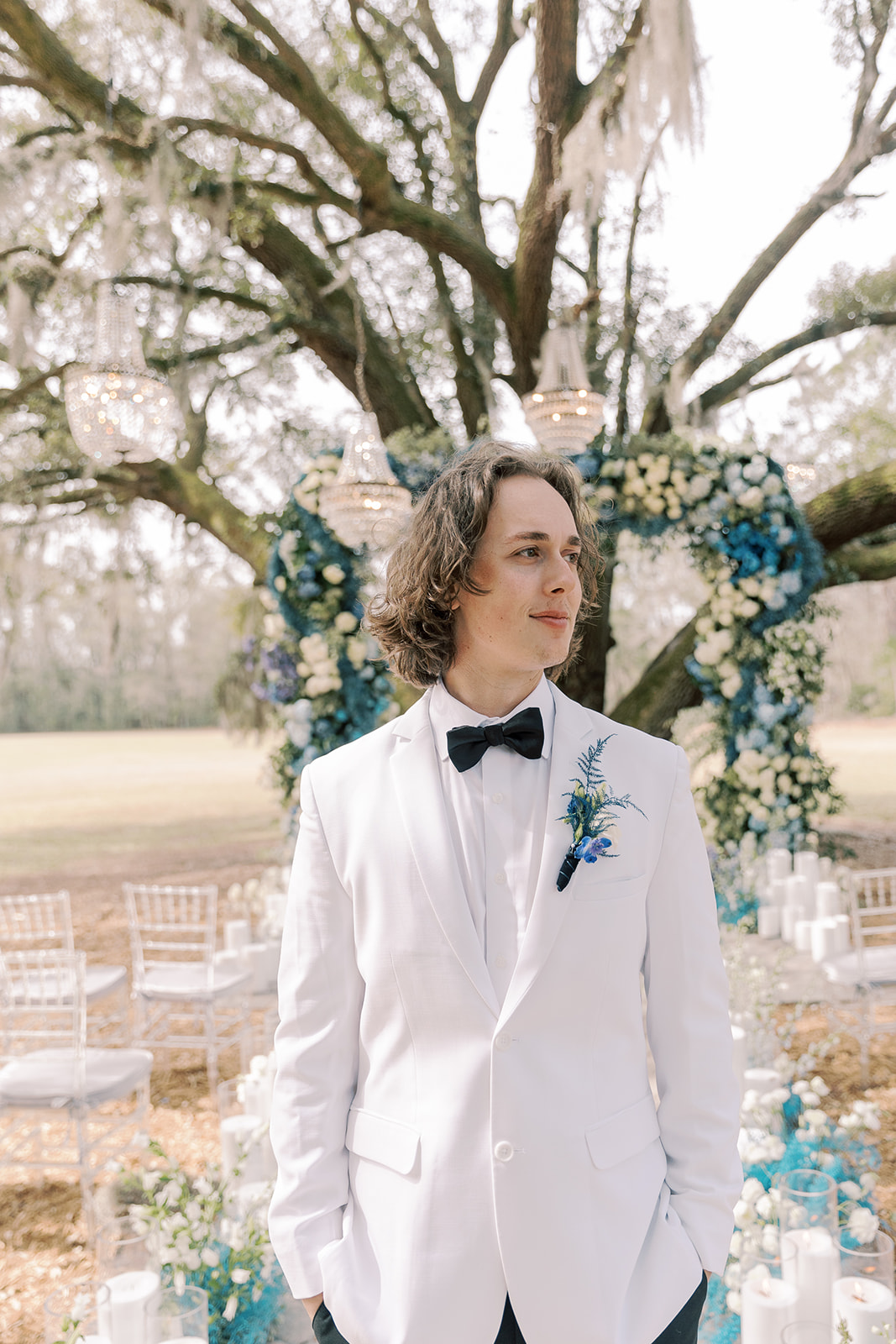 A man in a white suit with a bow tie stands outdoors in front of a floral wedding arch under a large tree.