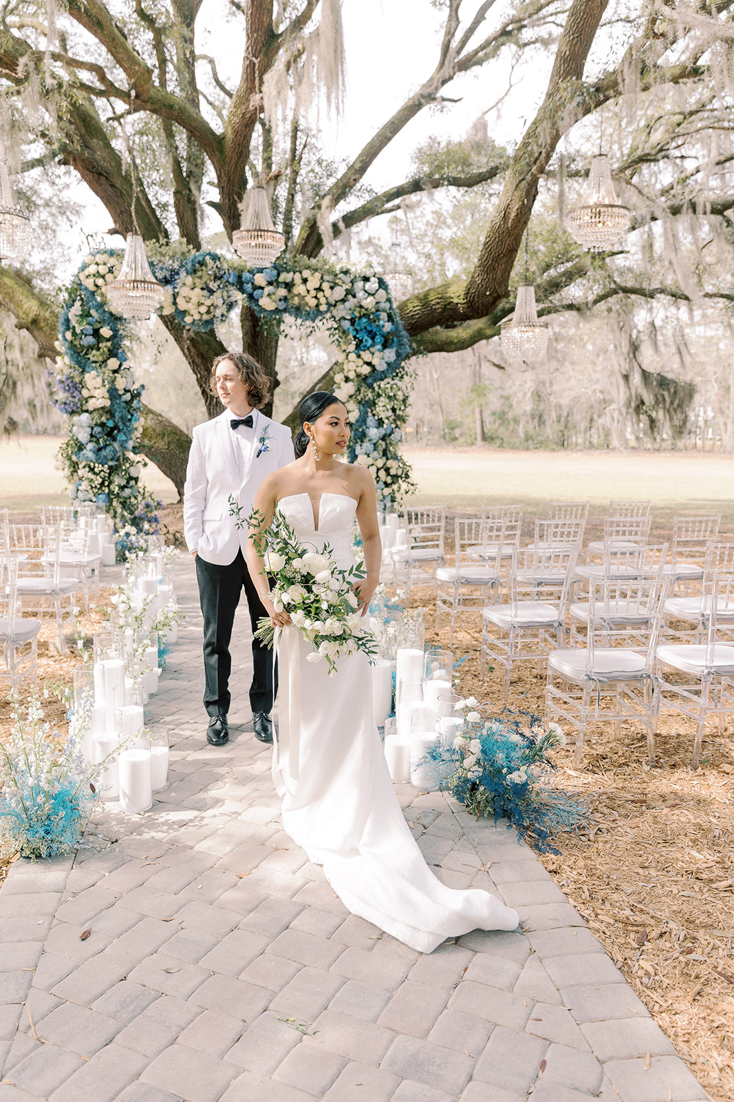 A bride and groom walk down an outdoor aisle decorated with flowers and white candles, under a large tree adorned with hanging lanterns and a floral arch at Hewitt Oaks venue