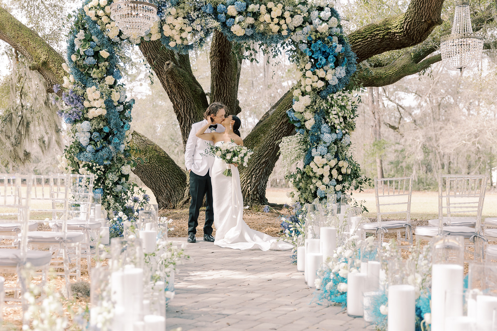 A bride and groom walk down an outdoor aisle decorated with flowers and white candles, under a large tree adorned with hanging lanterns and a floral arch.