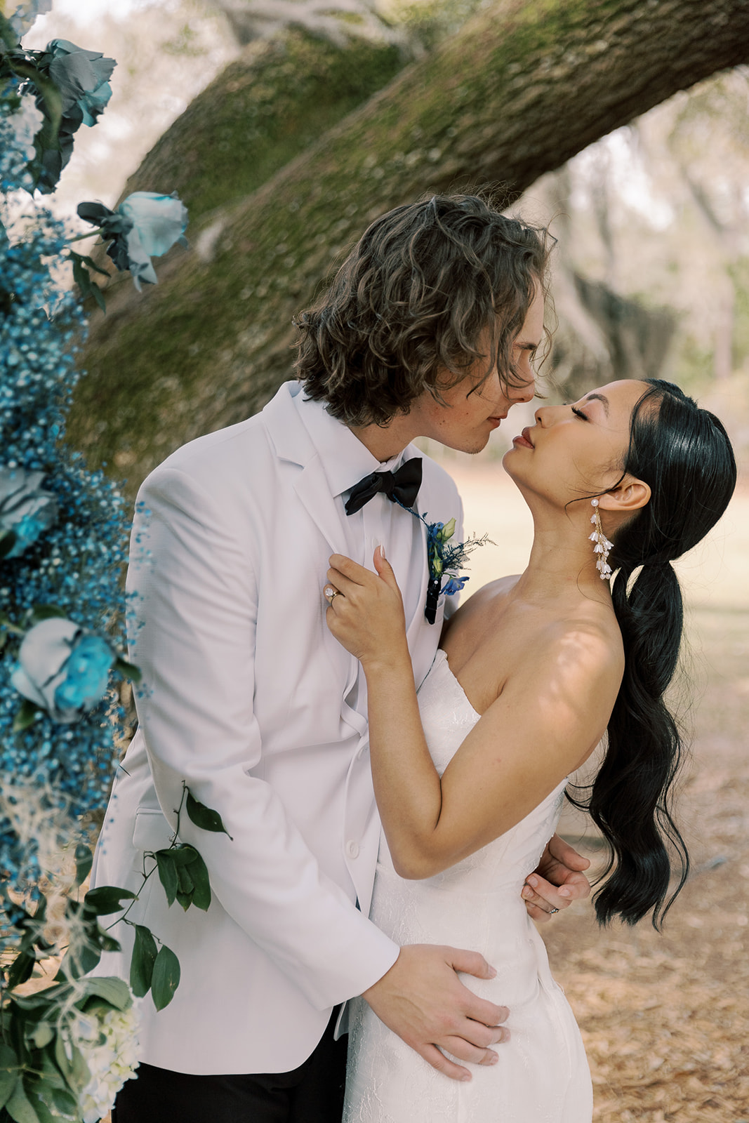 A couple kisses outdoors beside a flower arrangement. The man wears a white suit; the woman is in a strapless wedding gown at Hewitt Oaks venue