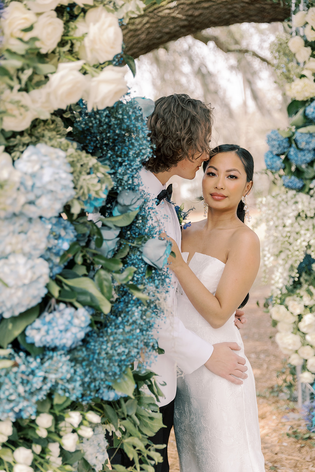 A couple kisses outdoors beside a flower arrangement. The man wears a white suit; the woman is in a strapless wedding gown at Hewitt Oaks venue