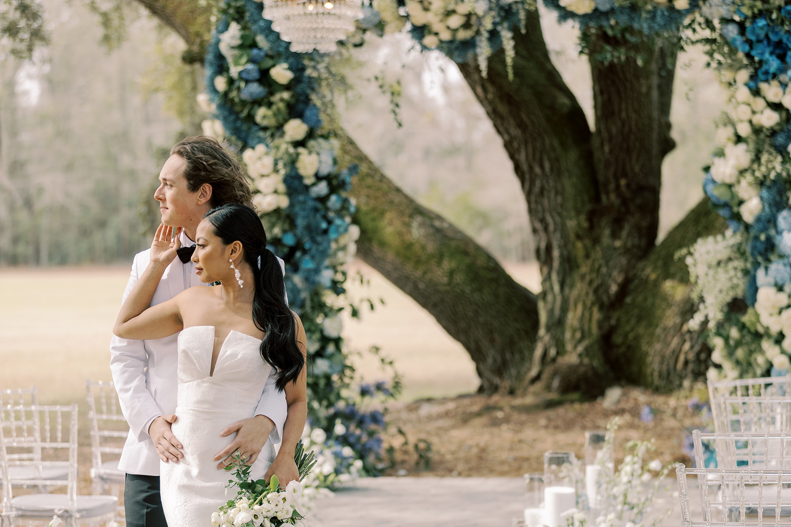 A bride and groom stand in front of a tree adorned with blue and white flowers. The groom rests his hand on the bride's shoulder as she holds a bouquet at Hewitt Oaks wedding venue