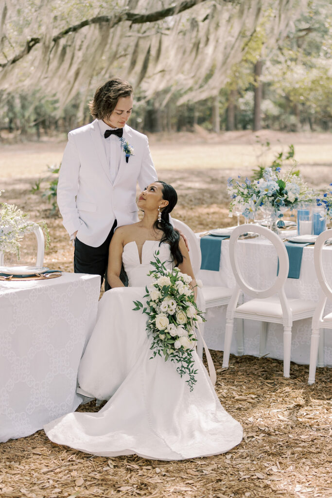 A bride, in a strapless white gown holding a bouquet, sits beside a groom in a white suit and black bowtie at an outdoor wedding reception table set with blue and white decorations at Hewitt Oaks wedding venue