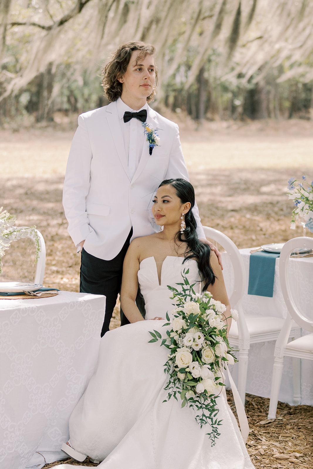 A bride, in a strapless white gown holding a bouquet, sits beside a groom in a white suit and black bowtie at an outdoor wedding reception table set with blue and white decorations at Hewitt Oaks wedding venue