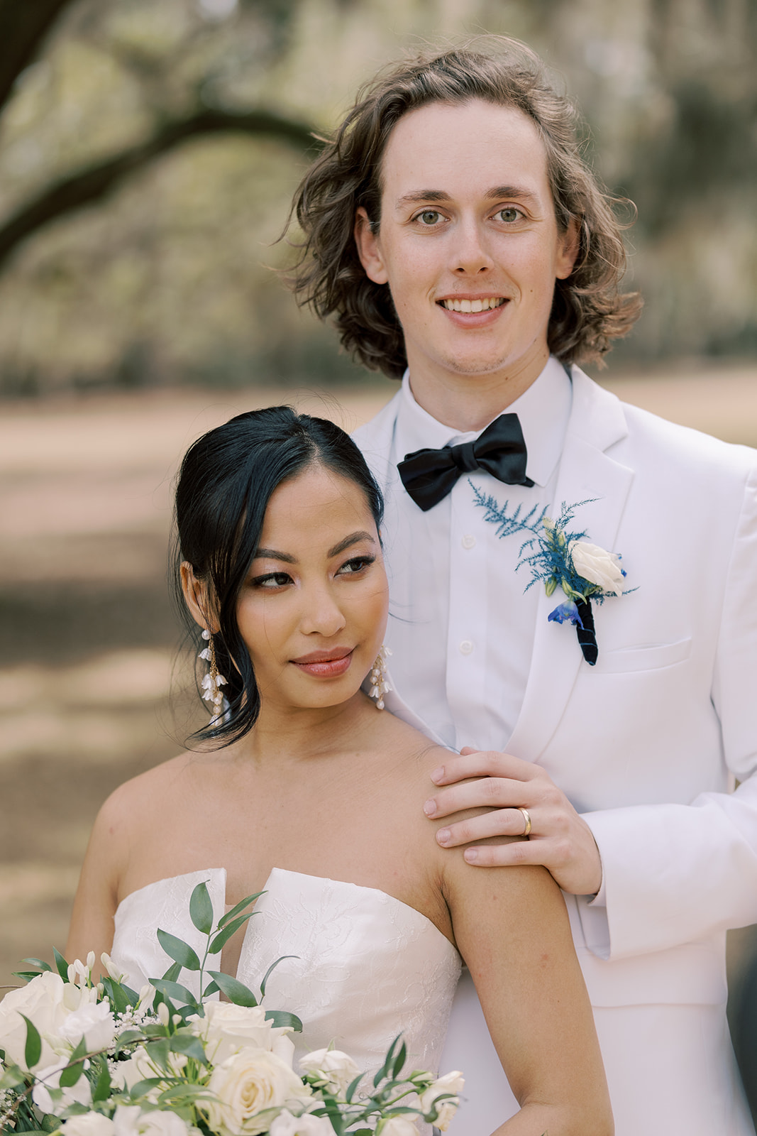 A bride and groom stand outdoors, the groom in a white tuxedo with a bowtie and the bride in a strapless wedding dress, holding a bouquet of white flowers.