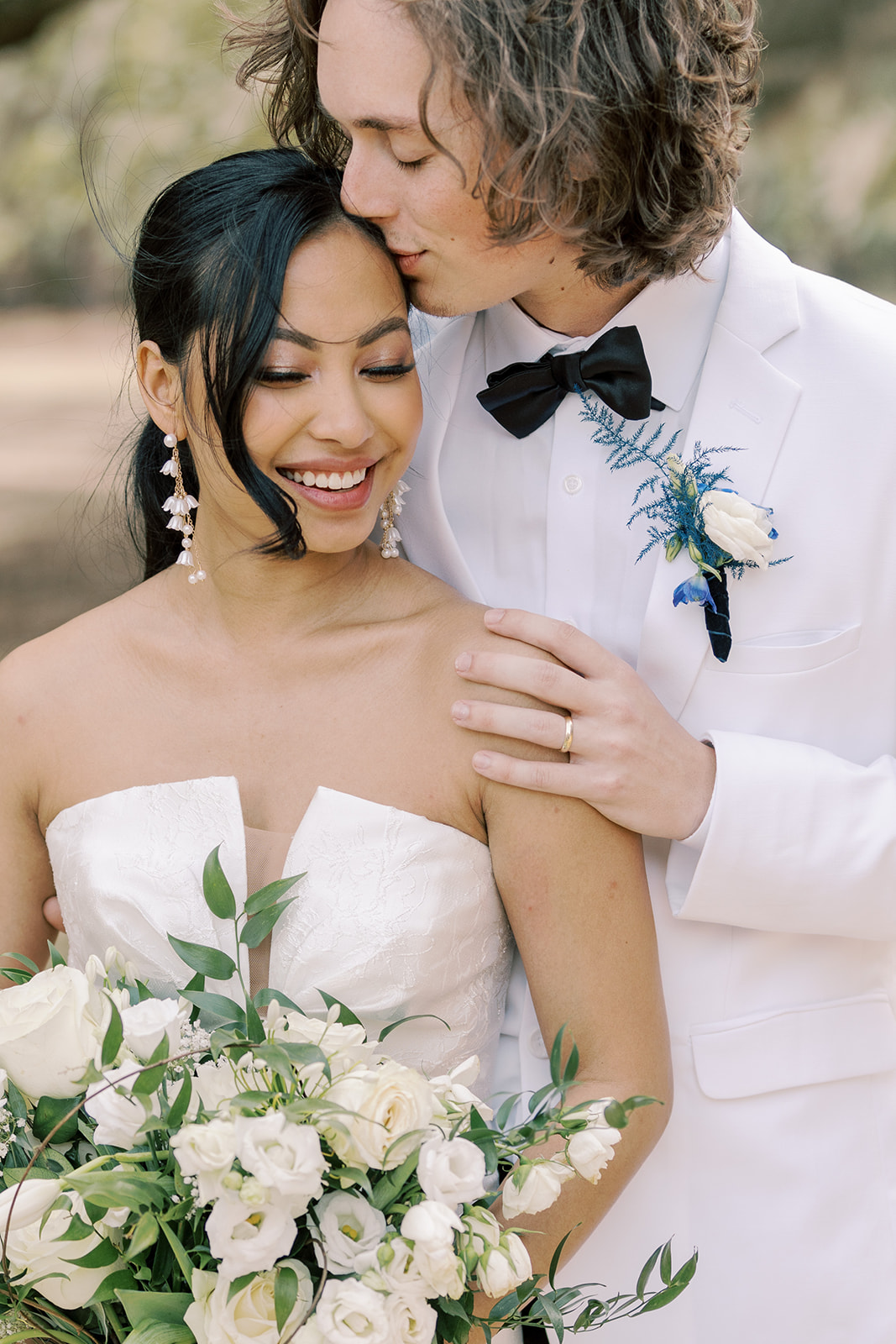 A bride and groom stand outdoors, the groom in a white tuxedo with a bowtie and the bride in a strapless wedding dress, holding a bouquet of white flowers.