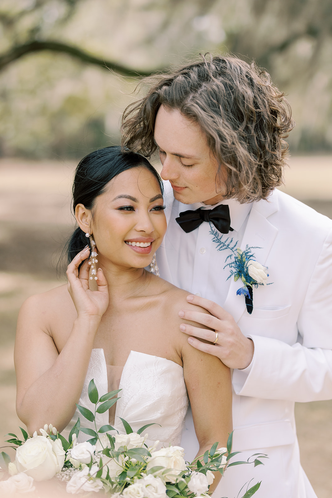 A bride and groom stand outdoors, the groom in a white tuxedo with a bowtie and the bride in a strapless wedding dress, holding a bouquet of white flowers.