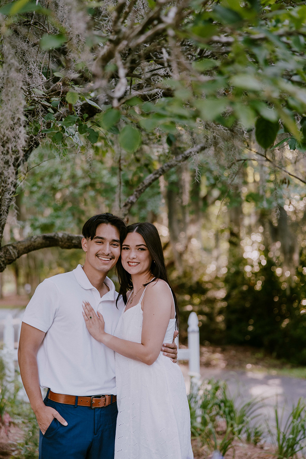 A smiling couple stands together in a lush outdoor setting. The man wears a white polo shirt and blue pants, while the woman is in a white sleeveless dress. They are surrounded by trees and greenery at Savannah botanical garden for an engagement session