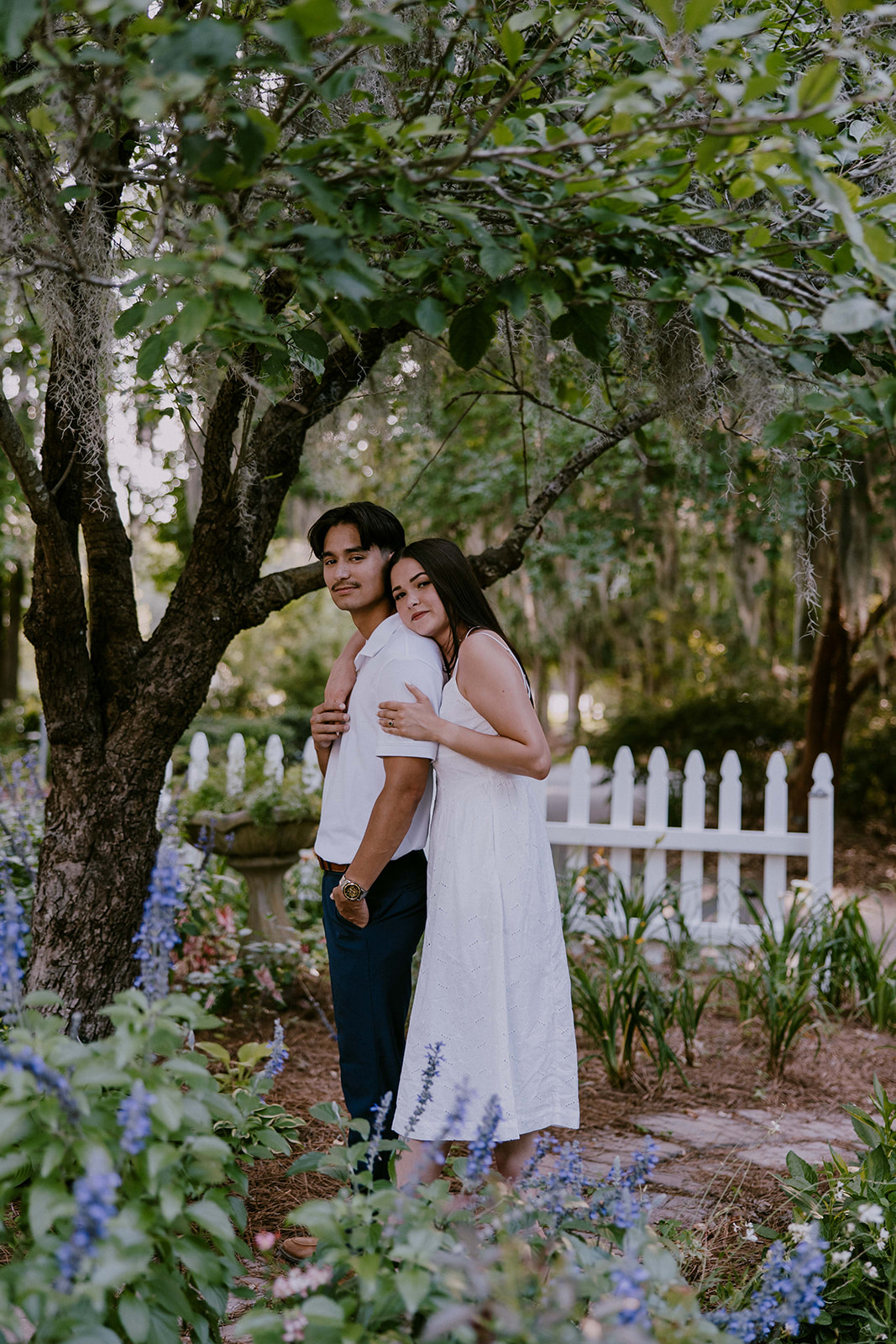 A smiling couple stands together in a lush outdoor setting. The man wears a white polo shirt and blue pants, while the woman is in a white sleeveless dress. They are surrounded by trees and greenery at Savannah botanical garden for an engagement session