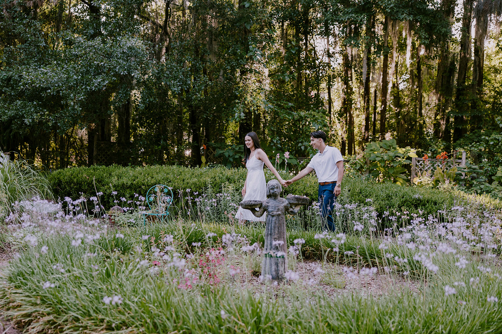 A smiling couple stands together in a lush outdoor setting. The man wears a white polo shirt and blue pants, while the woman is in a white sleeveless dress. They are surrounded by trees and greenery at Savannah botanical garden for an engagement session