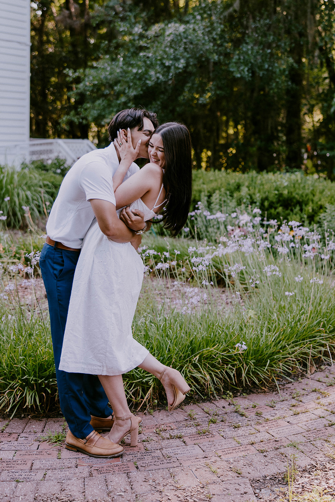 A smiling couple stands together in a lush outdoor setting. The man wears a white polo shirt and blue pants, while the woman is in a white sleeveless dress. They are surrounded by trees and greenery at Savannah botanical garden for an engagement session