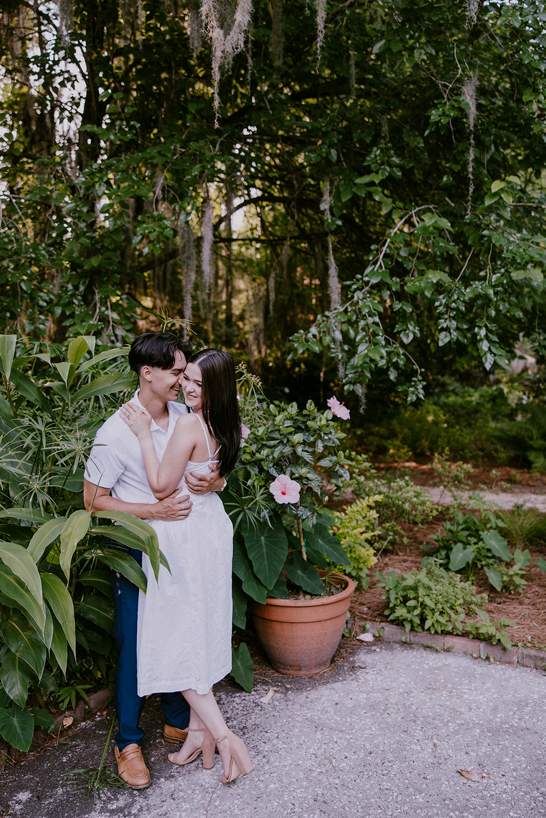 A smiling couple stands together in a lush outdoor setting. The man wears a white polo shirt and blue pants, while the woman is in a white sleeveless dress. They are surrounded by trees and greenery at Savannah botanical garden for an engagement session