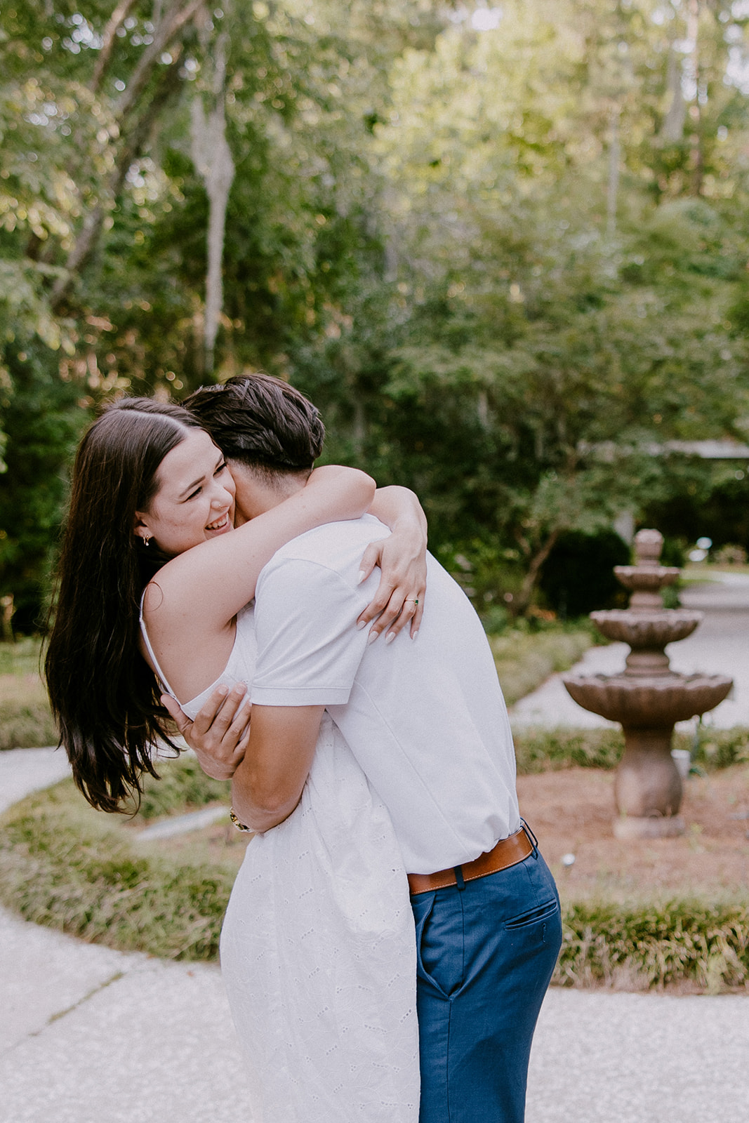 A smiling couple stands together in a lush outdoor setting. The man wears a white polo shirt and blue pants, while the woman is in a white sleeveless dress. They are surrounded by trees and greenery at Savannah botanical garden for an engagement session