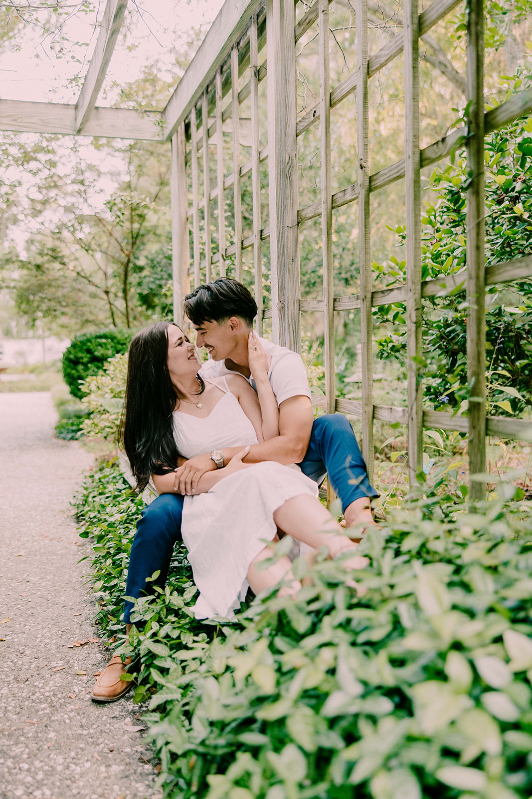 A smiling couple stands together in a lush outdoor setting. The man wears a white polo shirt and blue pants, while the woman is in a white sleeveless dress. They are surrounded by trees and greenery at Savannah botanical garden for an engagement session