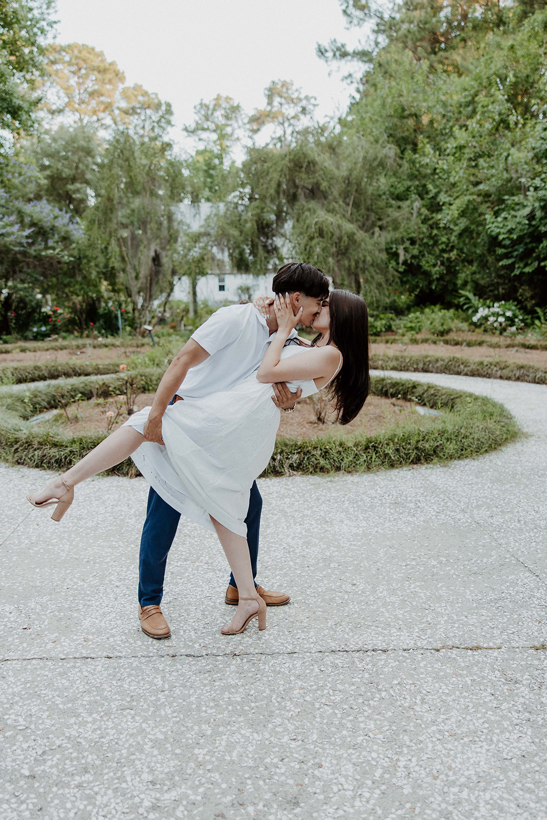 A smiling couple stands together in a lush outdoor setting. The man wears a white polo shirt and blue pants, while the woman is in a white sleeveless dress. They are surrounded by trees and greenery at Savannah botanical garden for an engagement session