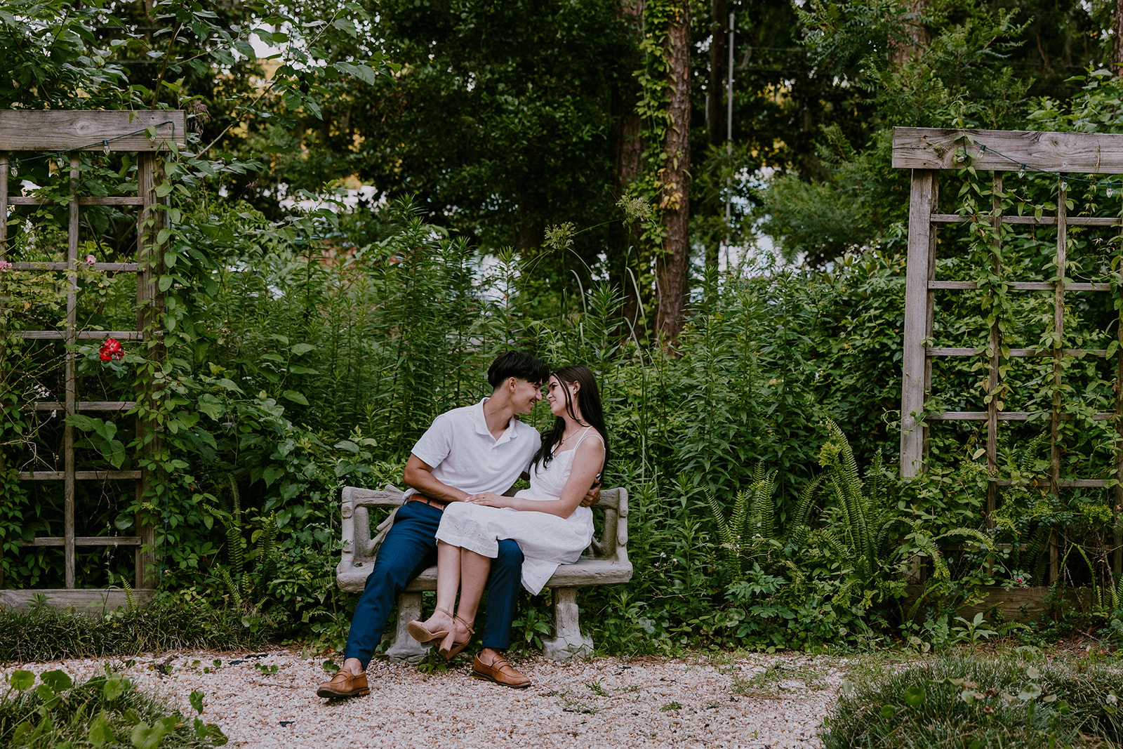 A smiling couple stands together in a lush outdoor setting. The man wears a white polo shirt and blue pants, while the woman is in a white sleeveless dress. They are surrounded by trees and greenery at Savannah botanical garden for an engagement session