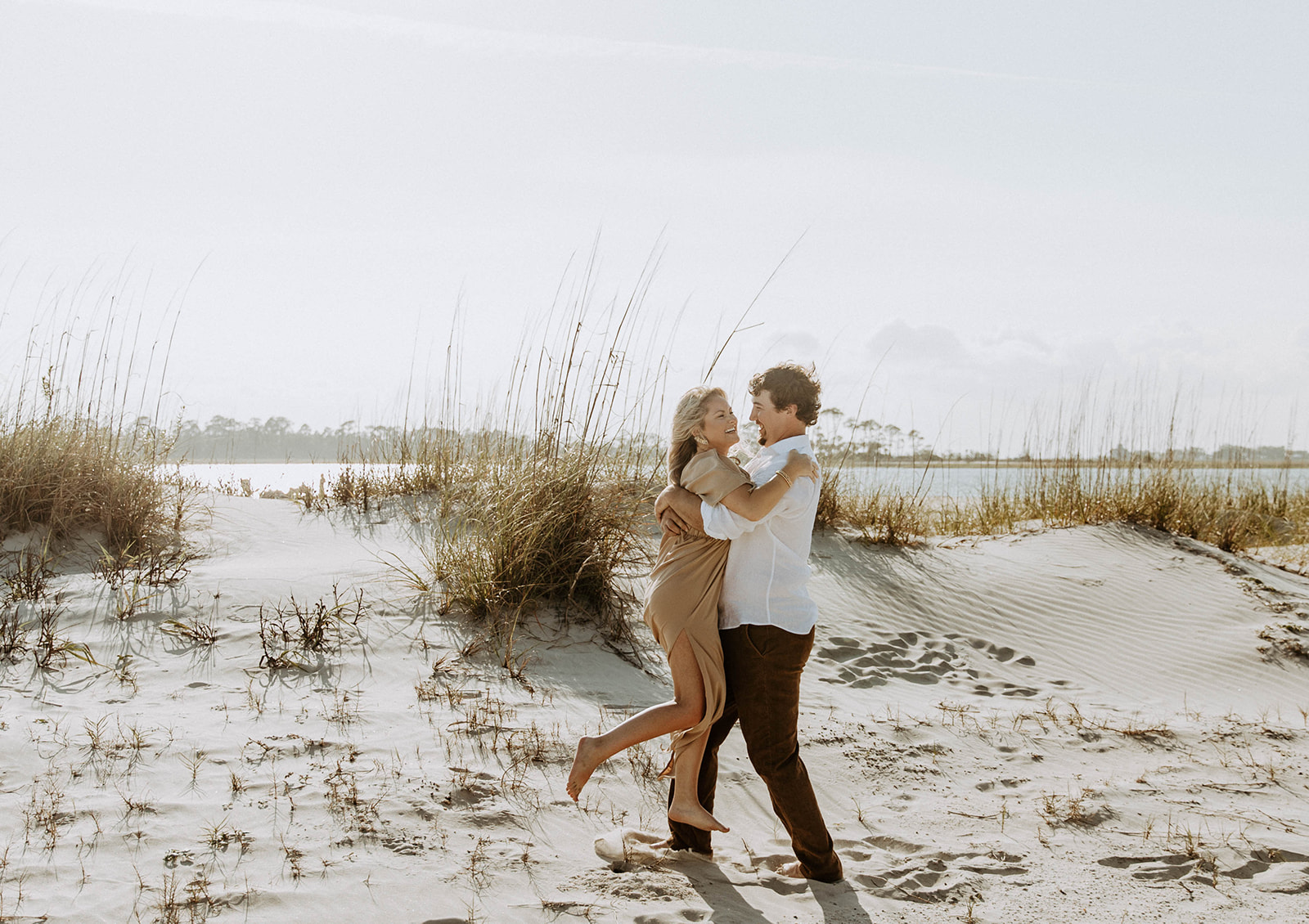 Couple joyfully hugging each other while standing on a sandy beach with tall grasses and a body of water in the background.