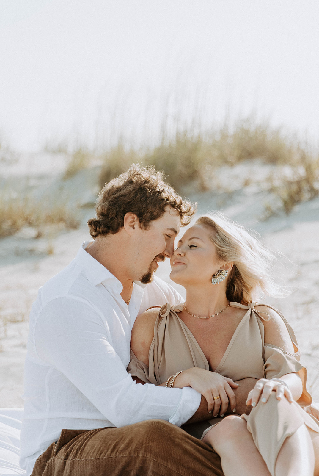 A man and a woman sit closely together on a sandy beach, looking at each other affectionately. The background features dunes and sparse vegetation.