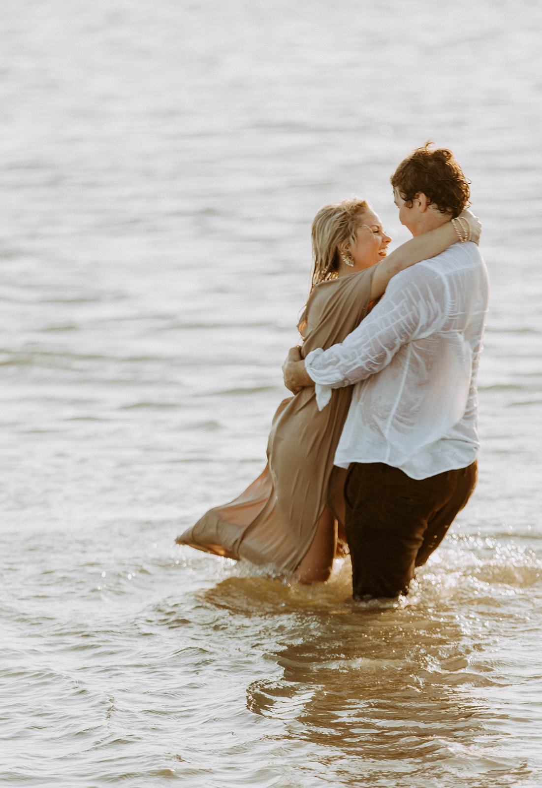 Two people stand in shallow water embracing each other. One wears a white shirt, and the other wears a light-colored dress.