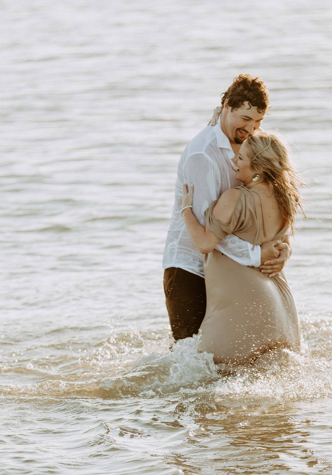 Two people stand in shallow water embracing each other. One wears a white shirt, and the other wears a light-colored dress.