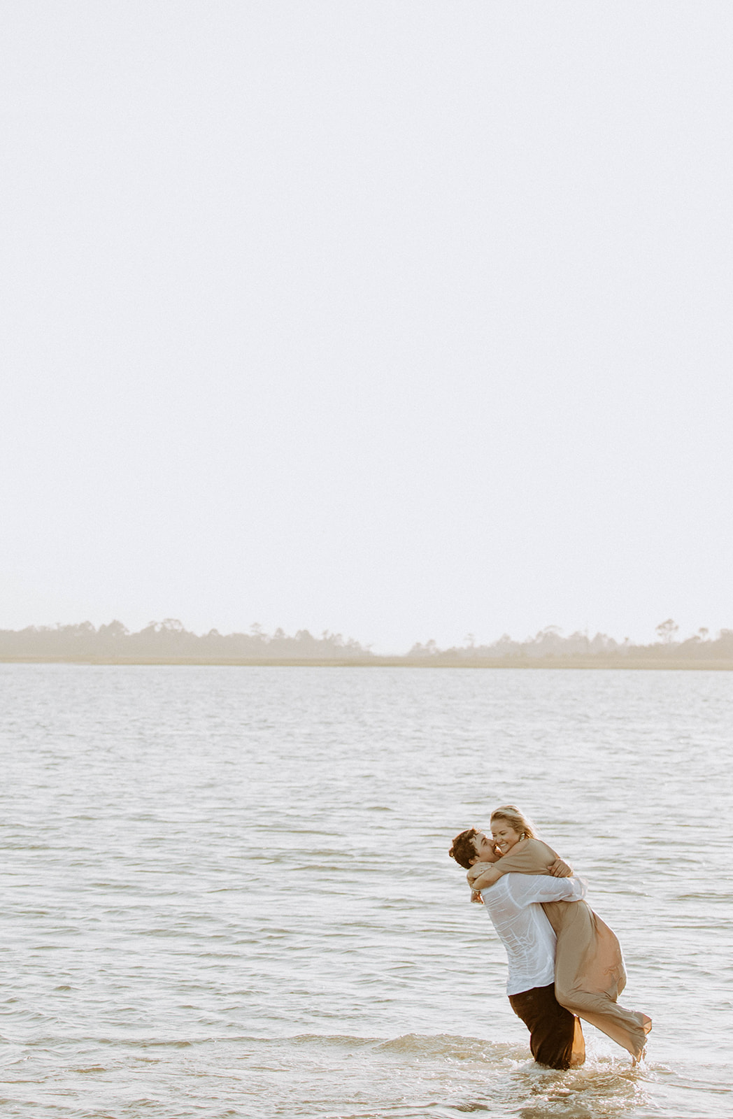 Two people stand in shallow water embracing each other. One wears a white shirt, and the other wears a light-colored dress.