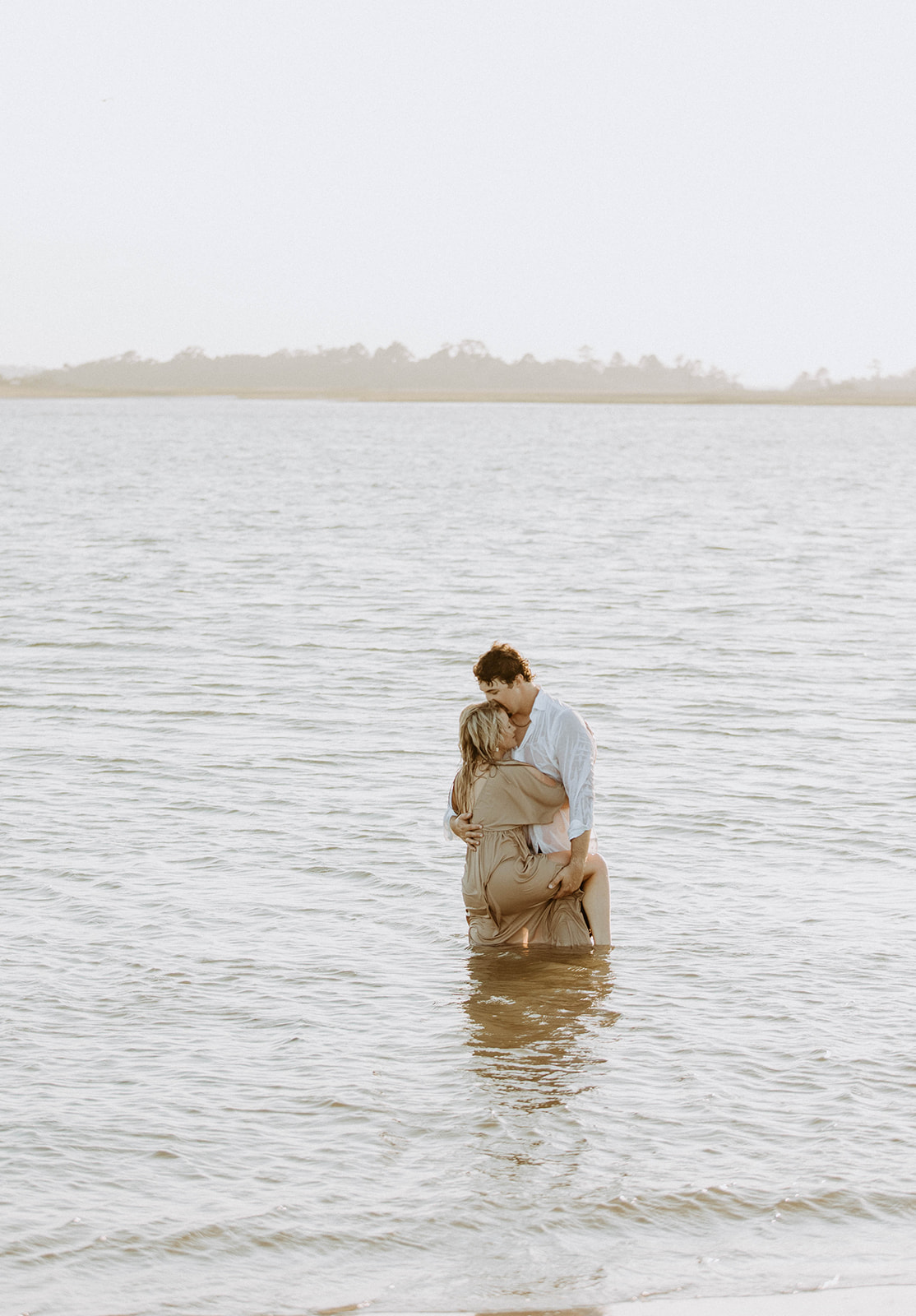 Two people stand in shallow water embracing each other. One wears a white shirt, and the other wears a light-colored dress.