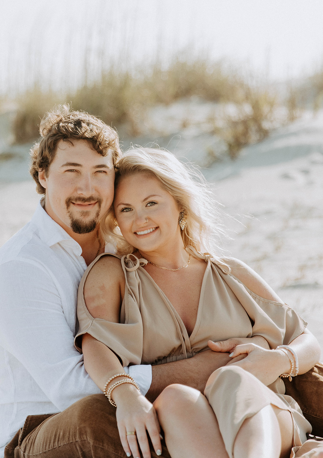 A man and a woman sit closely together on a sandy beach, looking at each other affectionately. The background features dunes and sparse vegetation.