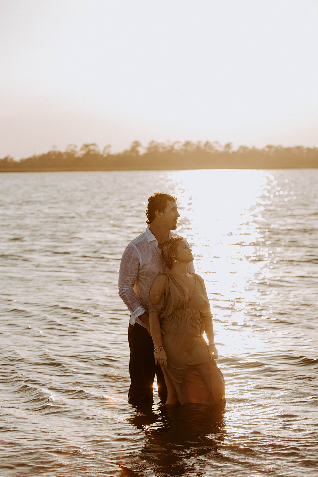 A couple standing together in shallow water at sunset, with the woman's hand resting on the man's arm.