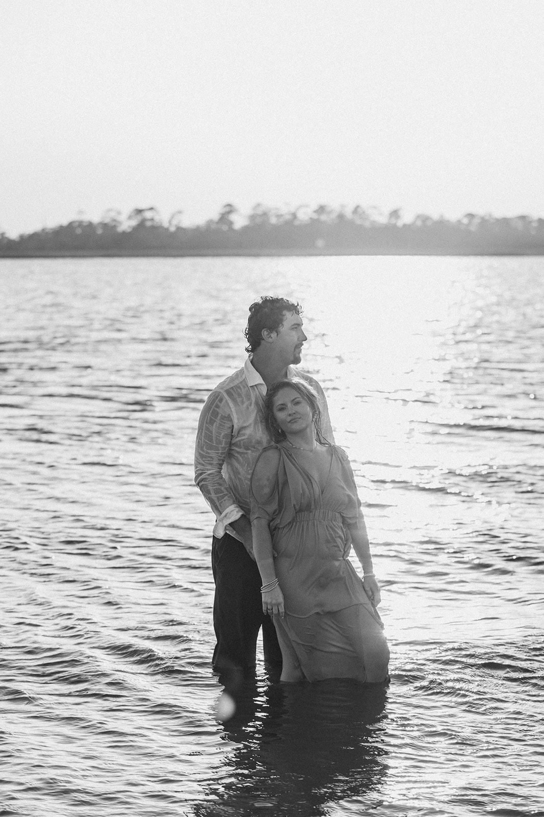 A couple stands in the water at sunset; the man stands behind the woman holding her arms as she leans back against him.