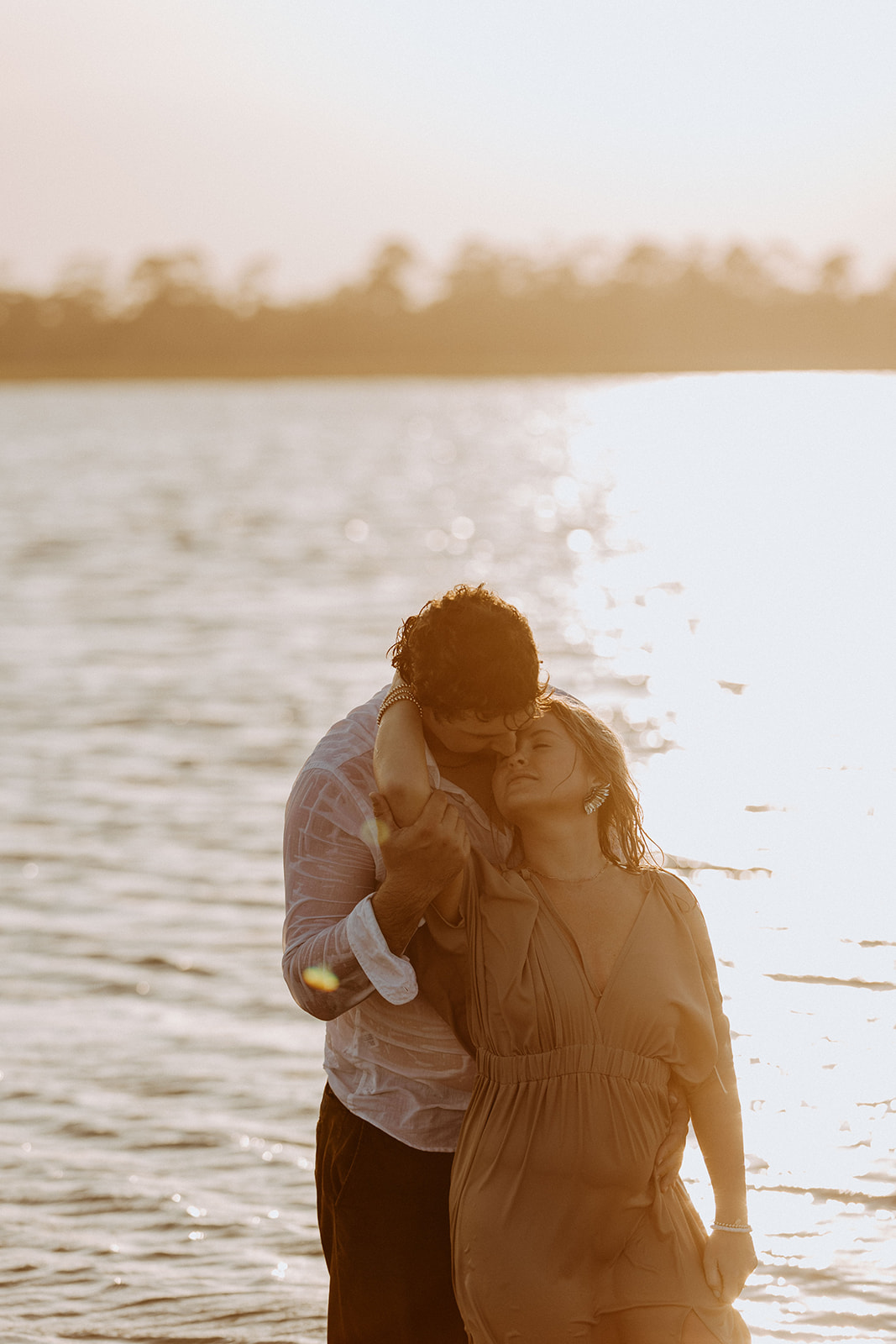 A couple stands in the water at sunset; the man stands behind the woman holding her arms as she leans back against him.