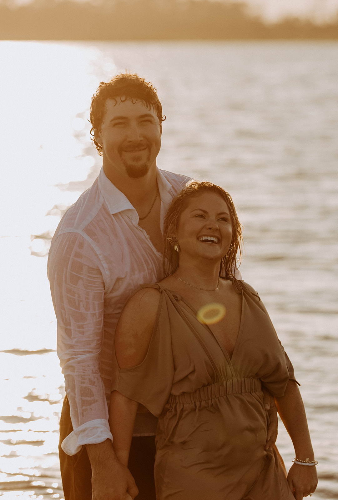 A couple stands in the water at sunset; the man stands behind the woman holding her arms as she leans back against him.