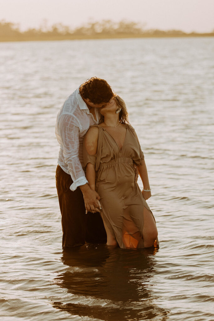 A couple stands in the water at sunset; the man stands behind the woman holding her arms as she leans back against him.