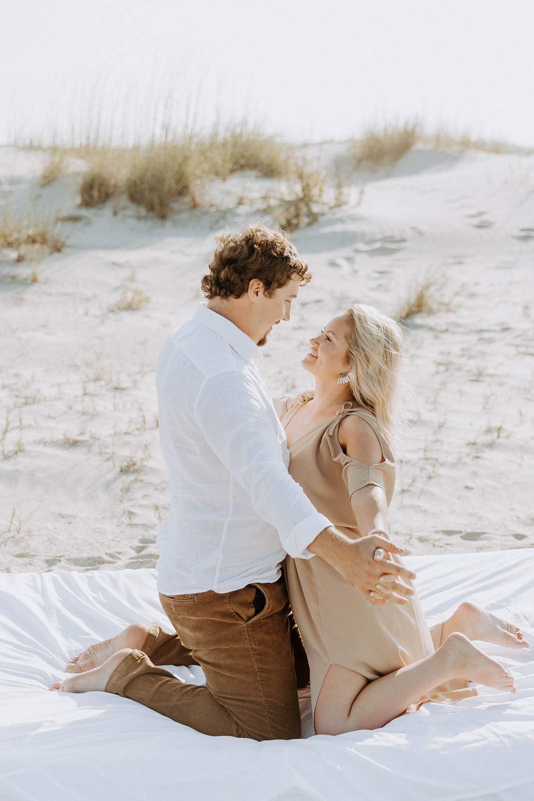 A man and a woman lie on a white blanket on a sandy beach, smiling and holding hands on Tybee Island