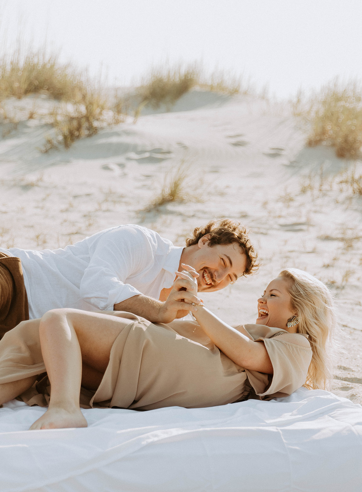 A man and a woman lie on a white blanket on a sandy beach, smiling and holding hands on Tybee Island