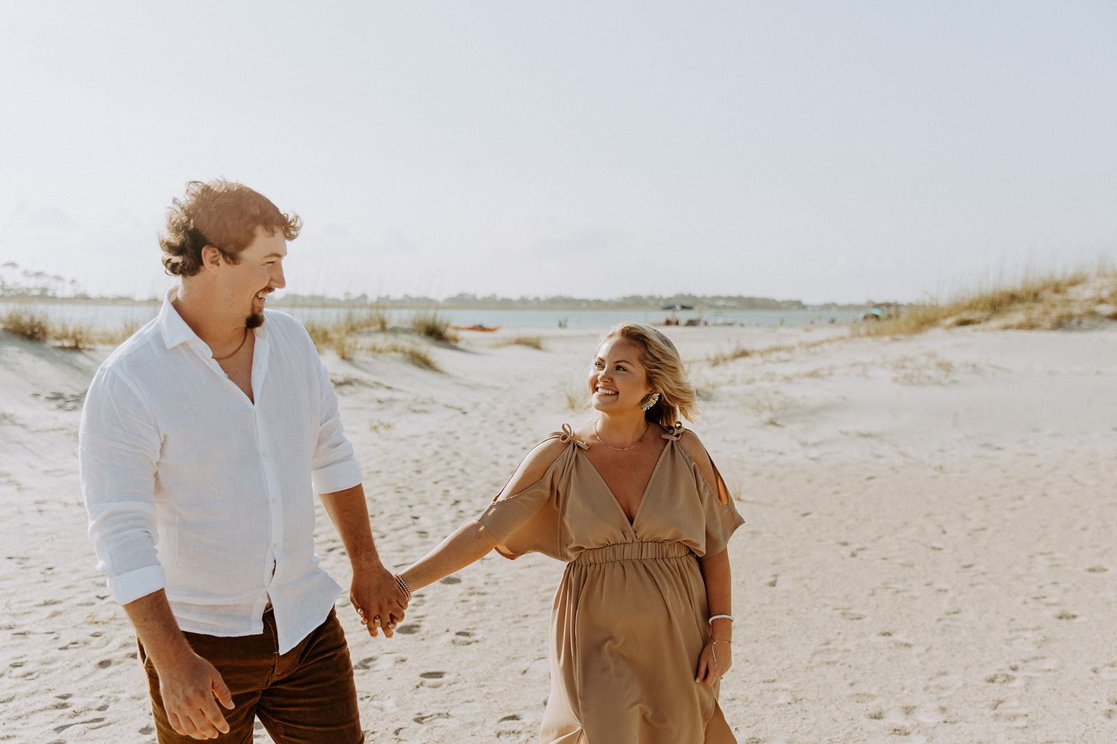 A couple walks hand in hand on a sandy beach under a clear sky on Tybee Island for their engagement photos