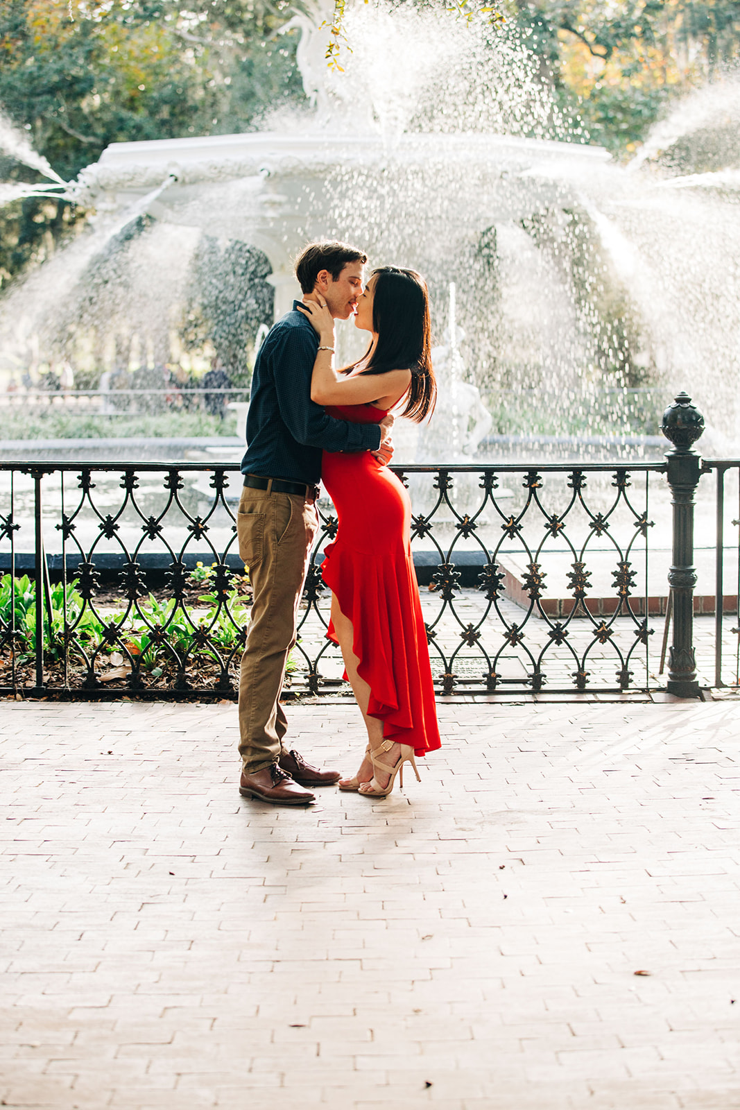 A couple is kissing on a sidewalk near a black iron fence. The man is wearing a blue shirt, and the woman is in a red dress at forysth park in savannah for their engagement