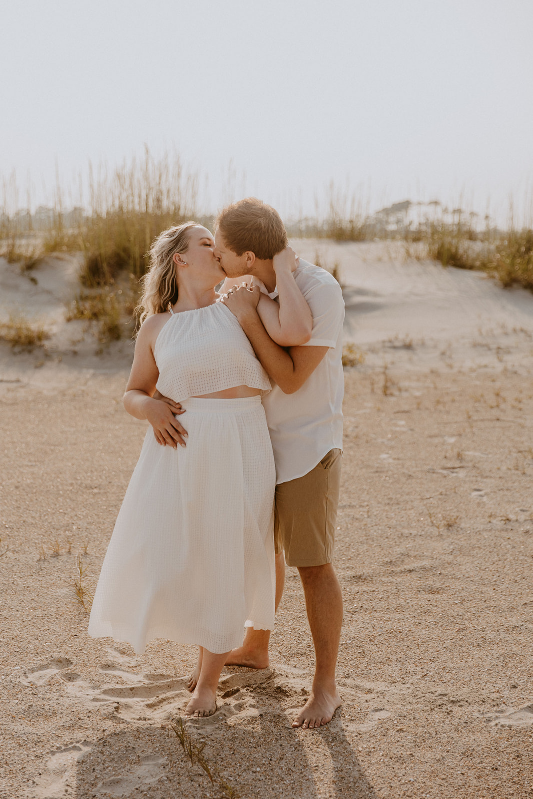 A couple, both dressed in light-colored clothing, embrace and kiss on a sandy beach with tall grass in the background for a savannah engagement on tybee island 