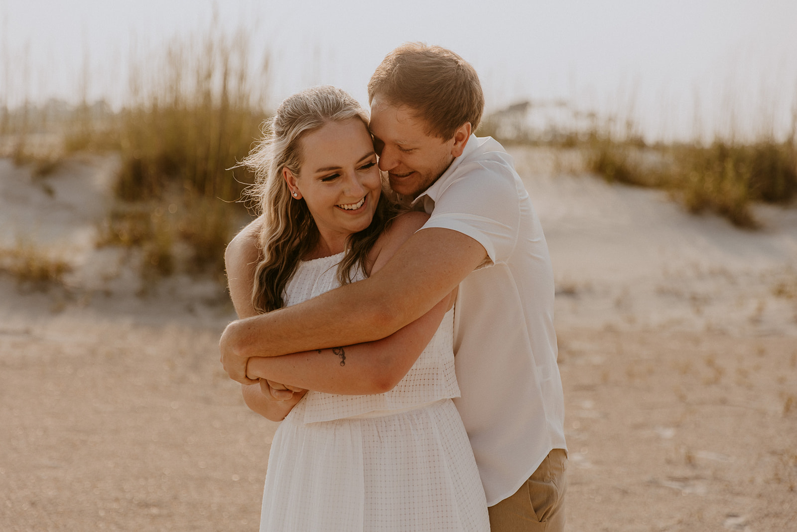 A couple, both dressed in light-colored clothing, embrace and kiss on a sandy beach with tall grass in the background for a savannah engagement on tybee island 