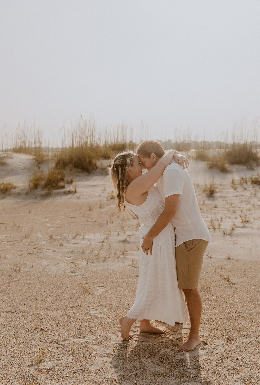 A couple, both dressed in light-colored clothing, embrace and kiss on a sandy beach with tall grass in the background for a savannah engagement on tybee island 