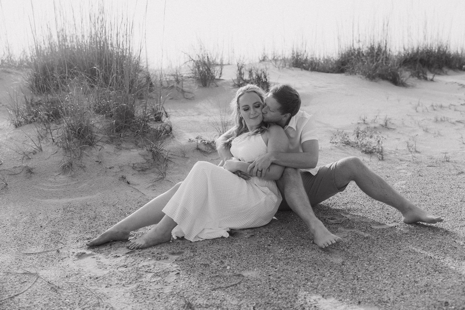 A couple, both dressed in light-colored clothing, embrace and kiss on a sandy beach with tall grass in the background for a savannah engagement on tybee island 