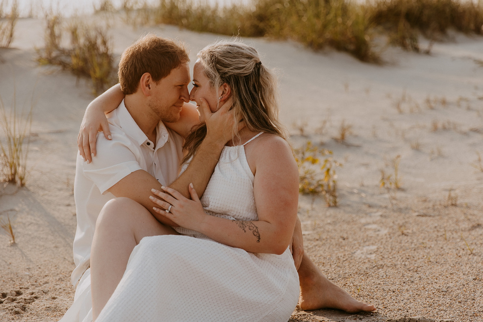 A couple, both dressed in light-colored clothing, embrace and kiss on a sandy beach with tall grass in the background for a savannah engagement on tybee island 