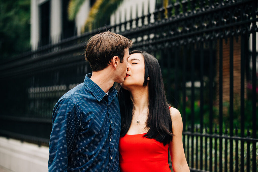 A couple is kissing on a sidewalk near a black iron fence. The man is wearing a blue shirt, and the woman is in a red dress at forysth park in savannah for their engagement