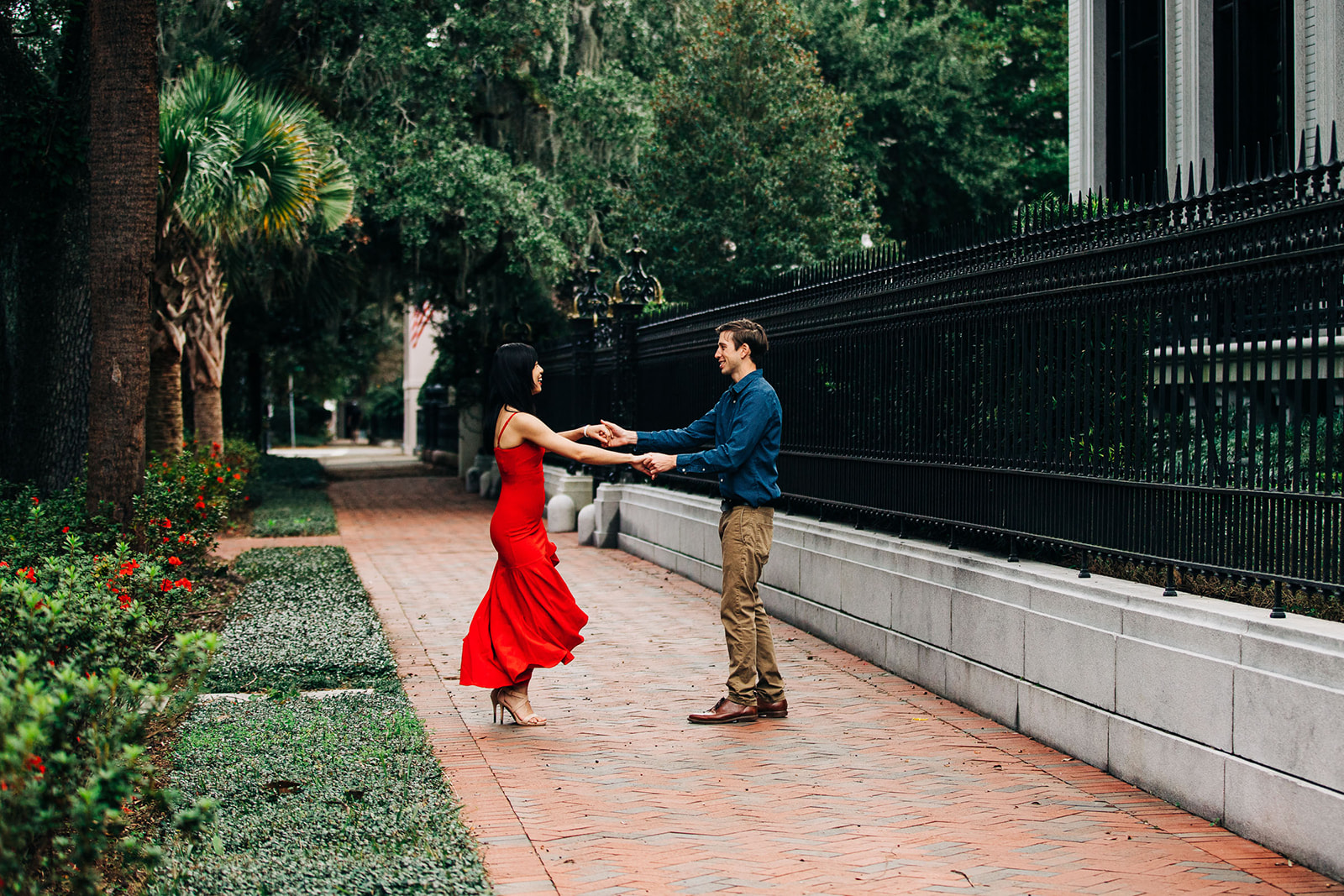 A couple is kissing on a sidewalk near a black iron fence. The man is wearing a blue shirt, and the woman is in a red dress at forysth park in savannah for their engagement